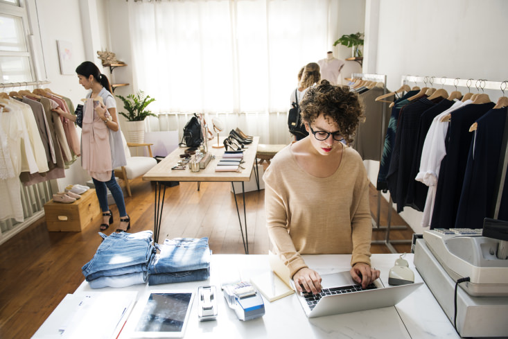 A woman small business owner working on her laptop while customers shop for clothes