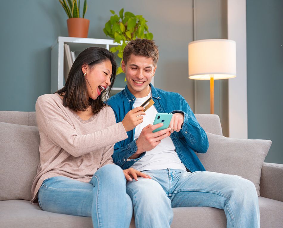 Two young adults sitting on a couch, smiling while making an online purchase with a smartphone and credit card.