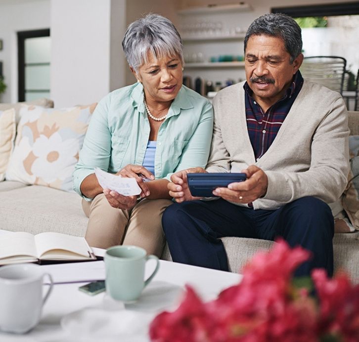Older couple sitting on a couch reviewing paperwork and using a tablet to manage their finances at home.